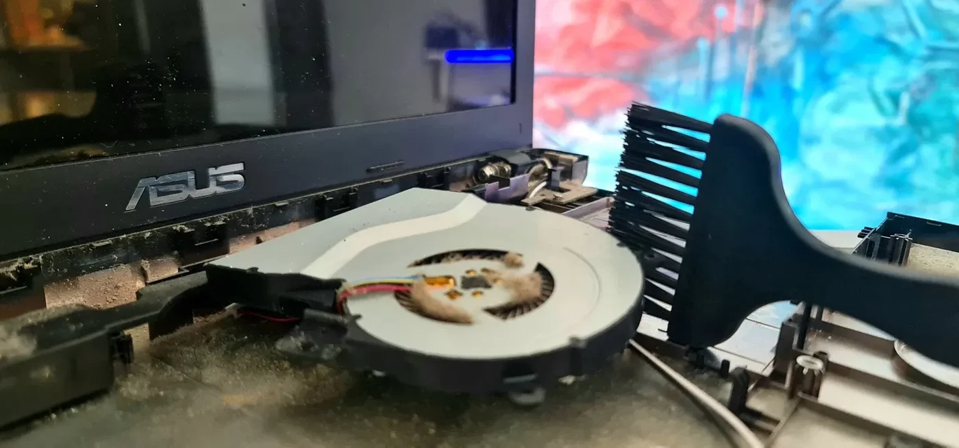 Notebook ASUS desarmada con la zona de la bisagra expuesta, el cooler con polvo y un pincel de limpieza